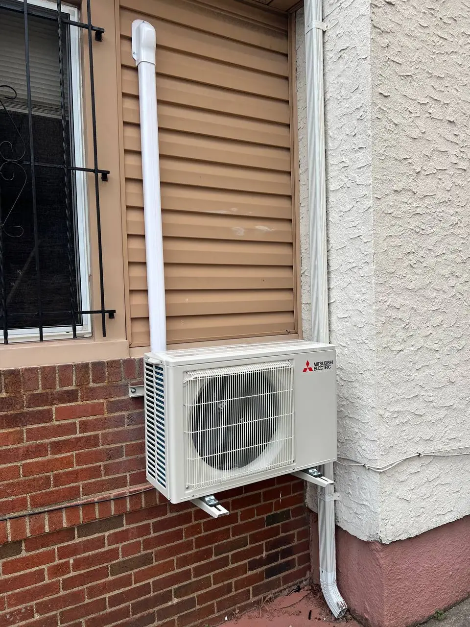 Compact condensing unit on wall brackets against a brick wall, refrigerant lines boxed in a white channel riser.