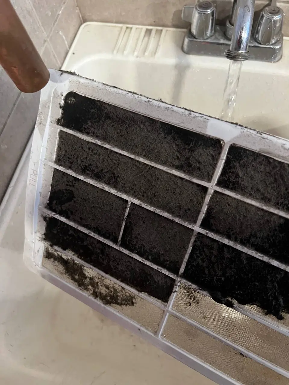 A mini-split filter held under a running faucet in a utility sink, its mesh blacked out with dust.