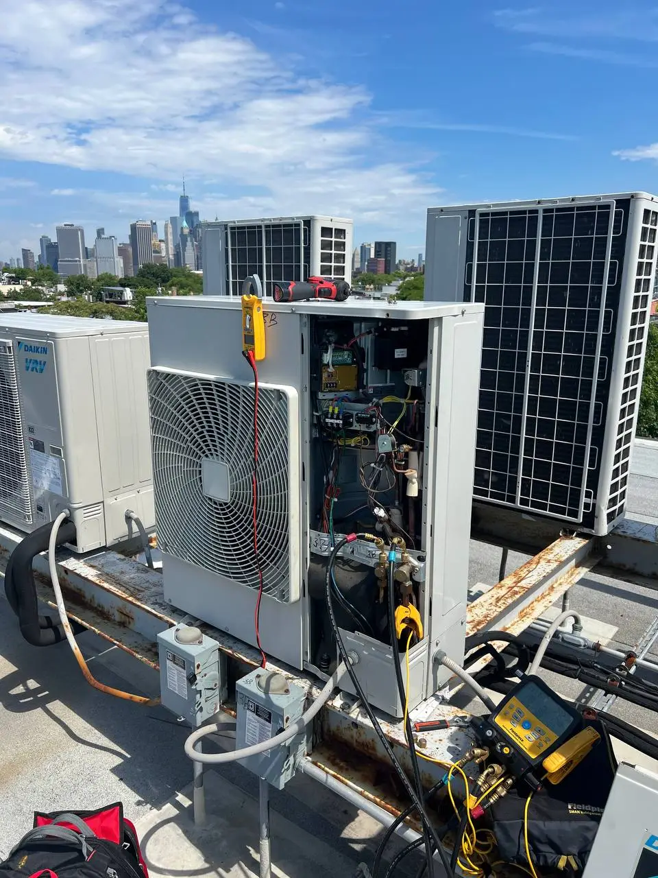 A rooftop condensing unit with its service panel open, a clamp meter and tools attached, and the Manhattan skyline behind it.