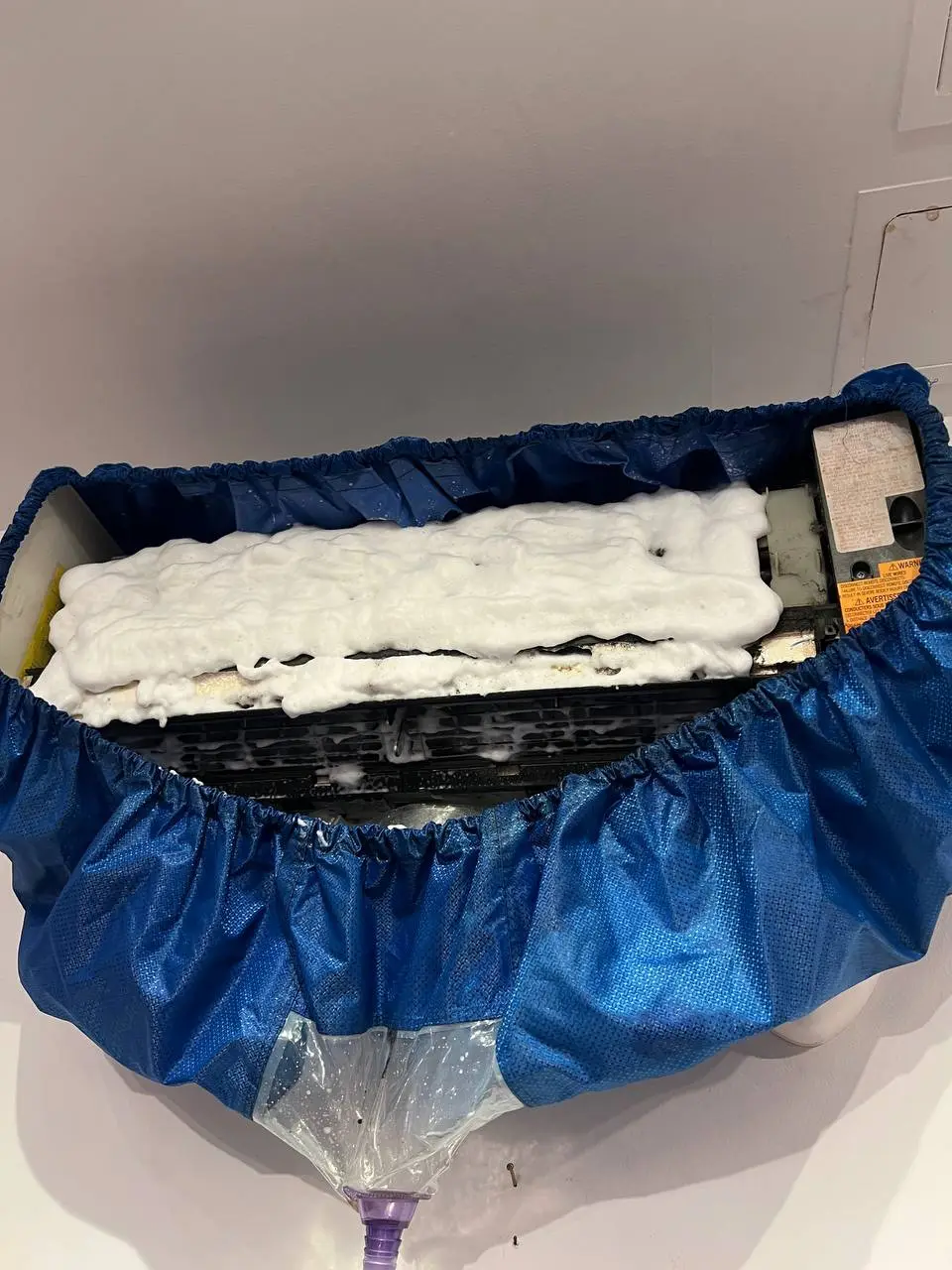 Indoor mini-split wrapped in a blue wash bag, its coil buried under white cleaning foam.